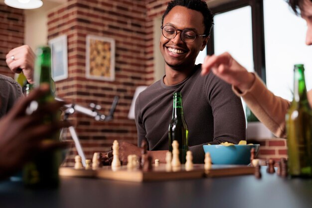 체스 게임 승리를 즐기면서 거실 테이블에 앉아 진심으로 웃는 남자. 여가 활동으로 휴식을 취하면서 다인종 경쟁적인 친구들과 함께 집에 앉아 있는 똑똑한 사람.