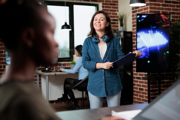 Smiling heartily office worker having clipboard with management plan standing in workspace while looking at camera. Creative agency confident employee in production department.