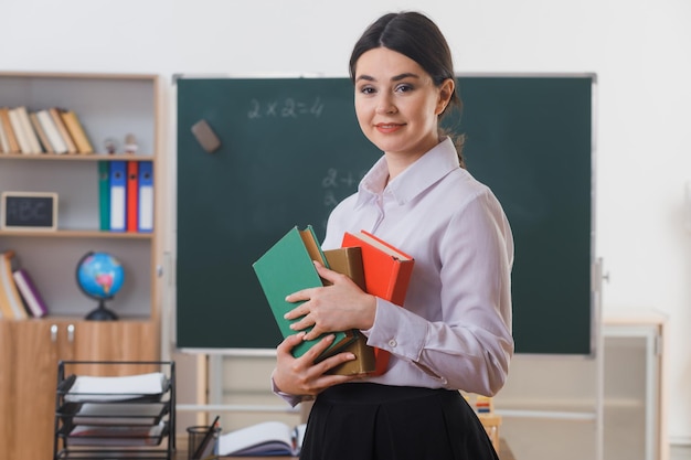 smiling holding books young teacher standing in front of blackboard in classroom