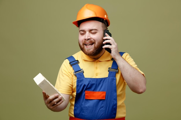 Smiling holding brick speaks on the phone young builder man in uniform isolated on green background