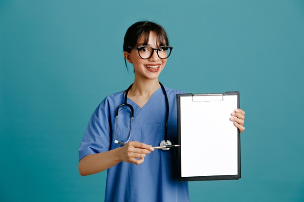 파란색 배경에 격리된 균일한 청진기를 입고 클립보드를 들고 웃고 있는 젊은 여성 의사