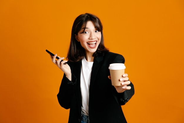 Улыбаясь, держа чашку кофе с телефоном молодая красивая женщина в черной куртке, изолированные на оранжевом фоне