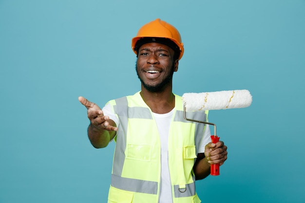 Улыбающийся протягивающий руку в камеру молодой афро-американский строитель в униформе, держащий роликовую щетку на синем фоне