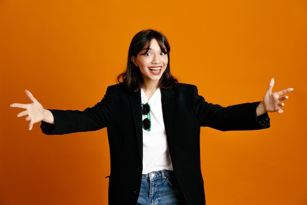 Smiling holding out hands at camera young beautiful female wearing black jacket isolated on orange background