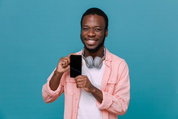 Free photo smiling holding phone young africanamerican guy wearing headphones on neck isolated on blue background