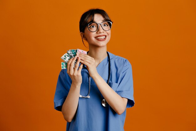 Smiling holding pills young female doctor wearing uniform fith stethoscope isolated on orange background