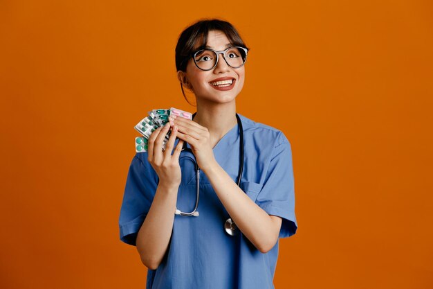Улыбаясь, держа таблетки молодая женщина-врач в униформе фито стетоскоп, изолированные на оранжевом фоне