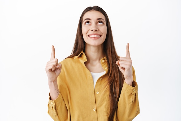 Smiling hopeful girl looking dreamy, pointing up and reading sign promo above, showing top advertisement or logo, standing over white wall in casual stylish clothes