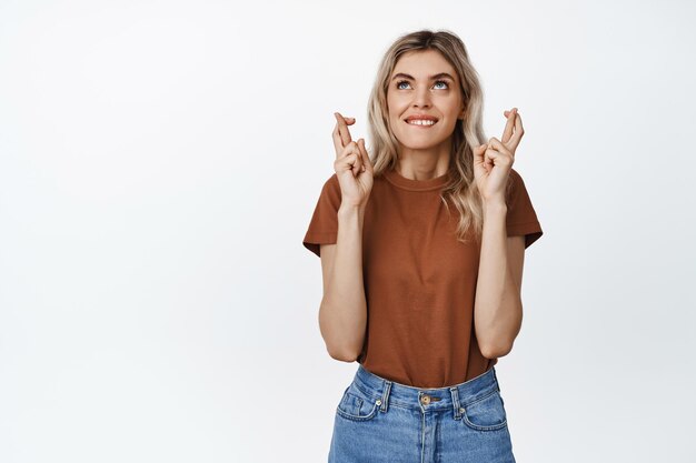 Smiling hopeful girl making wish hoping to get something with crossed fingers standing in casual outfit over white background