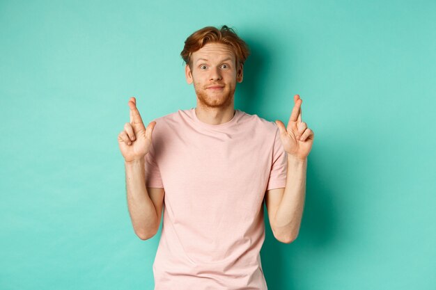 Smiling hopeful man with red hair making a wish, cross fingers for good luck and expecting something good, standing over turquoise background