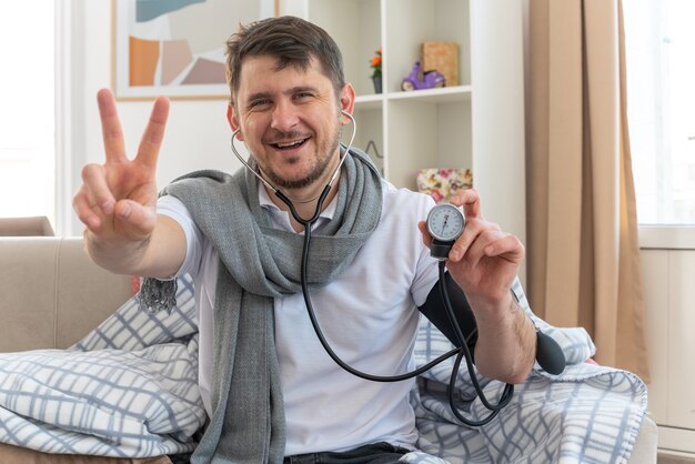 smiling ill man with scarf around neck measuring his pressure with sphygmomanometer and gesturing victory sign sitting on couch at living room