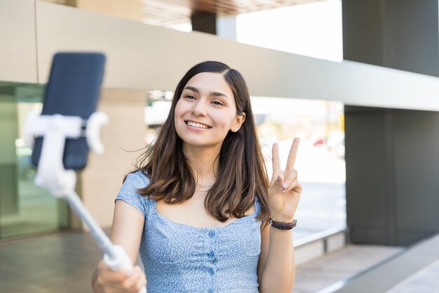 Smiling influencer gesturing victory sign while recording video on smartphone