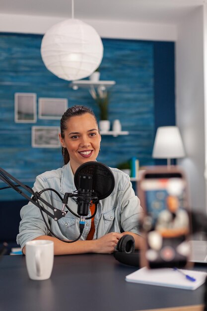 Smiling influencer streaming vlog in front of recording mobile phone. Content creator in home studio filming video for social media. Woman broadcasting podcast with microphone and smartphone.