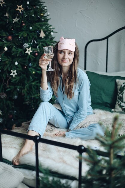 Улыбающаяся веселая женщина в синей пижаме и спальной маске, сидящая на кровати с бокалом шампанского. Рождество.