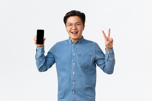 스마트폰 화면과 완두콩을 보여주는 안경과 교정기에 웃고 있는 즐거운 아시아 남성 프리랜서 학생...