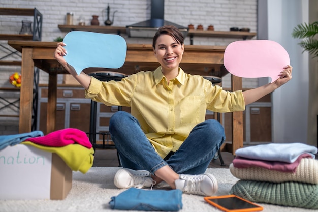 Smiling joyful dark-haired beautiful young woman sitting with her legs crossed among piles of clothes