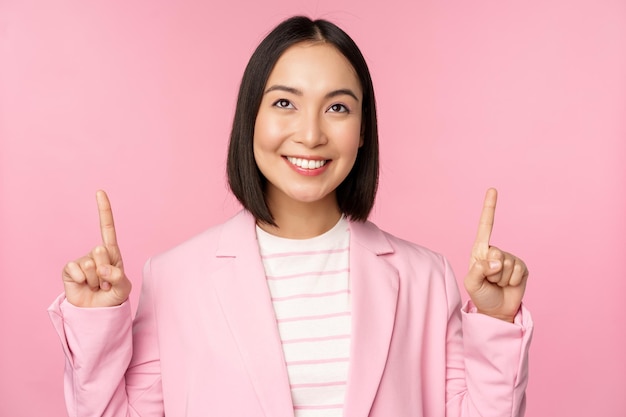 Smiling korean businesswoman pointing fingers up showing advertisement banner or logo on top standing in suit over pink background