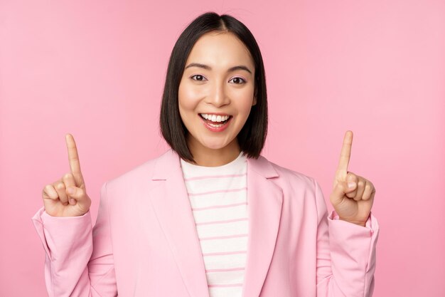 Smiling korean businesswoman pointing fingers up showing advertisement banner or logo on top standing in suit over pink background