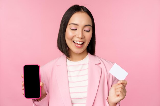 Smiling korean businesswoman in suit showing mobile phone screen credit card showing online banking application interface pink background