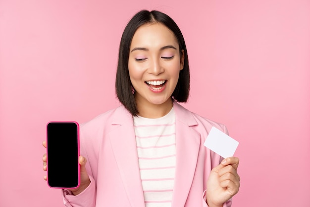 オンラインバンキングアプリケーションインターフェイスピンクの背景を示す携帯電話の画面のクレジットカードを示すスーツで笑顔の韓国の実業家