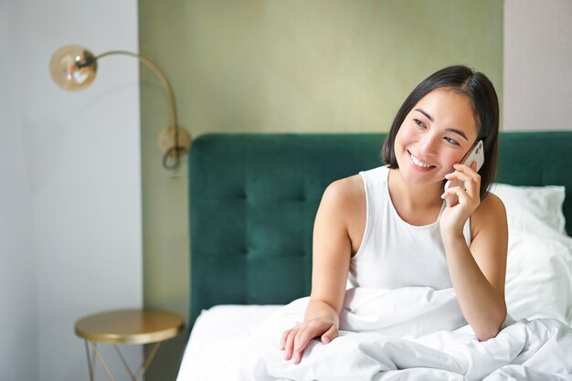 Smiling korean girl in bed talks on mobile phone making a phone call lazy morning as asian woman ord