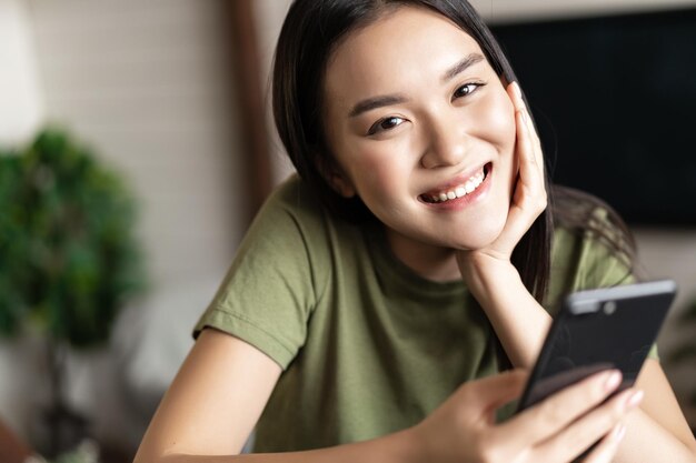 Smiling korean girl looking happy holding smartphone and sitting in living room at home lifestyle co...