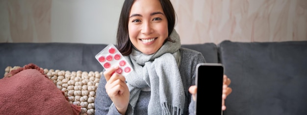 Free photo smiling korean girl shows smartphone screen medication in hands feeling sick and staying at home