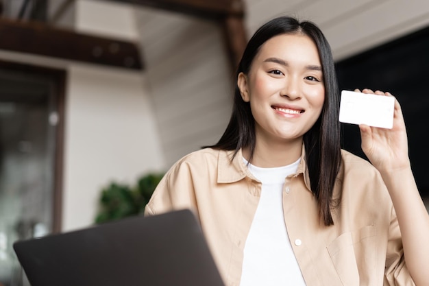 Smiling korean girl sitting at home with laptop showing credit card