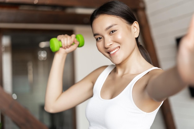 Smiling korean girl taking selfie with dumbbell, workout at home during pandemic, wearing activewear