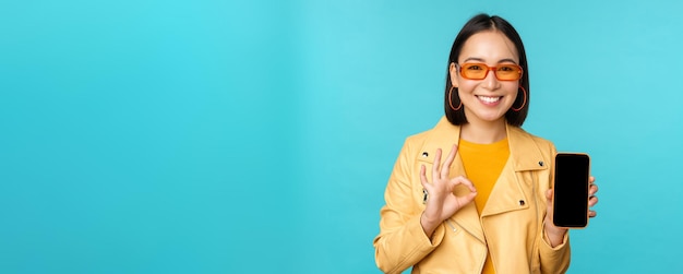 Smiling korean woman showing mobile phone app interface smartphone application recommending on cellphone standing over blue background