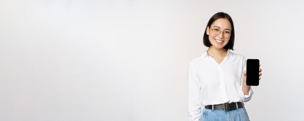 Free photo smiling korean woman showing smartphone screen demonstrating mobile application standing over white background