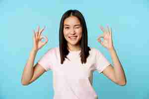 Free photo smiling korean woman winking showing okay signs recommending company or store standing in tshirt ove...