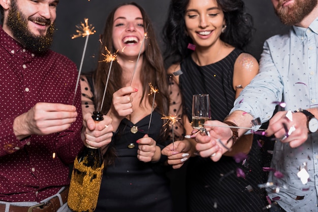 Smiling ladies and guys holding burning Bengal lights, bottle and glass of drink