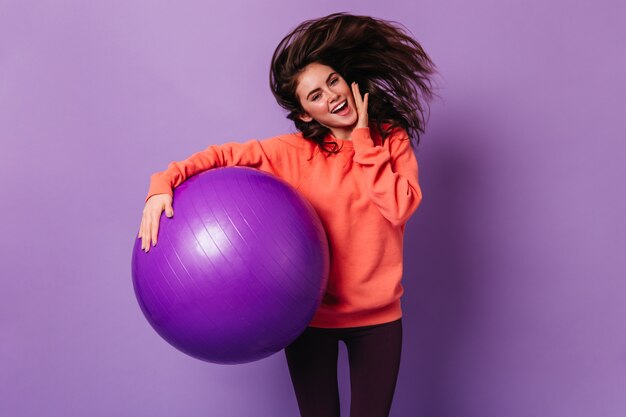 밝은 운동복과 어두운 레깅스에 웃는 아가씨가 보라색 벽에 점프하여 fitball을 들고