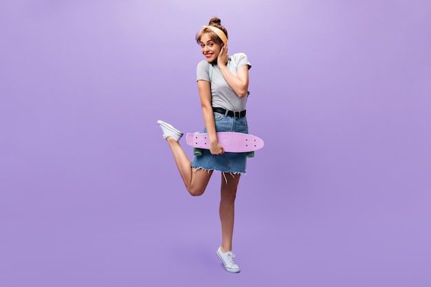 Smiling lady in denim outfit poses with longboard. Funny young woman in grey shirt and trendy skirt with black belt looking into camera.