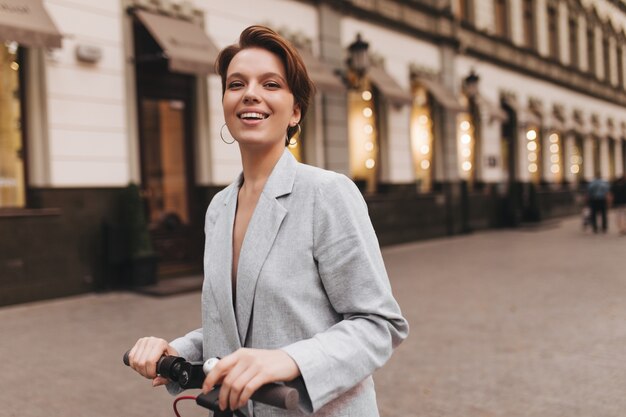 Smiling lady in grey outfit riding scooter. Cheerful young short-haired woman in stylish oversize jacket smiles sincerely and poses outside