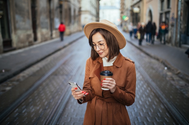 Smiling lady has a videocall and drinks coffee while walking outdoors in the city