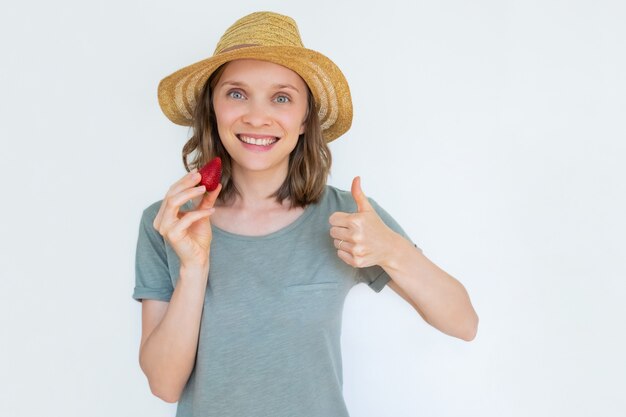 Smiling lady in hat holding ripe strawberry with thumb up