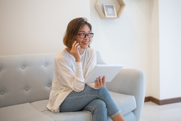 Smiling lady holding tablet and talking on phone at home