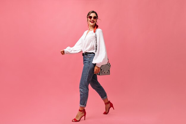 Smiling lady in jeans, white blouse walking on pink background.  Fashionable woman in red sunglasses steps on isolated backdrop.