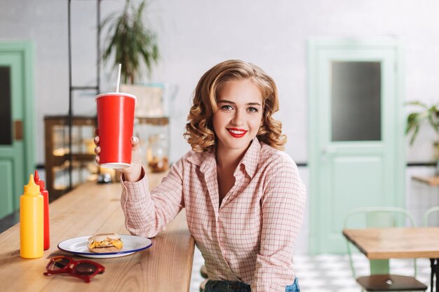 Smiling lady in shirt sitting at the bar counter with soda water in hand and burger near and happily looking in  camera while spending time in cafe