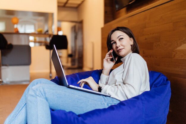 Smiling lady using mobile phone and laptop on bag chair in creative office