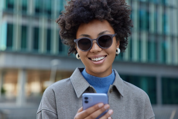 Smiling lady with afro hair bowses mobile phone while standing on city street being in good mood wears sunglasses and coat surfs social networks connected to wireless internet