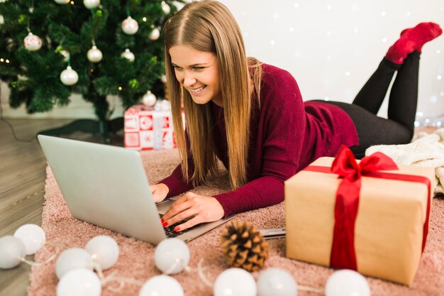 Smiling lady with laptop near gift boxes, snag, fairy lights and Christmas tree