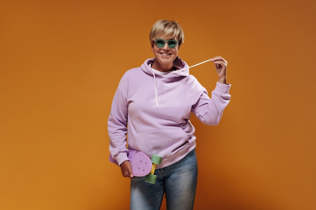 Smiling lady with short hair and modern glasses in pink sweatshirt and cool jeans posing with modern skateboard on orange backdrop. 