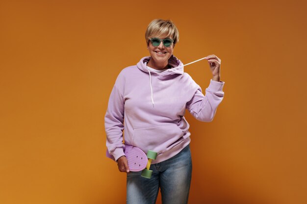Улыбающаяся дама с короткими волосами и в современных очках в розовой толстовке и крутых джинсах позирует с современным скейтбордом на оранжевом фоне.