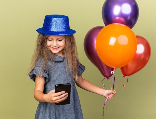 smiling little blonde girl with blue party hat holding helium balloons and looking at phone isolated on olive green wall with copy space