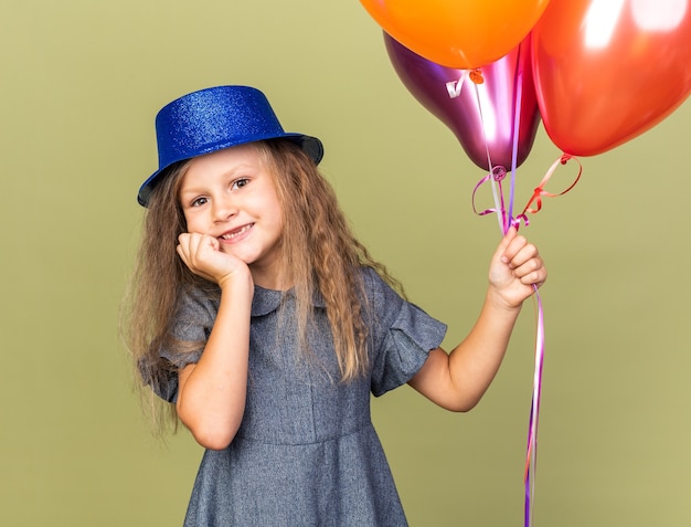 smiling little blonde girl with blue party hat holding helium balloons and putting hand on chin isolated on olive green wall with copy space