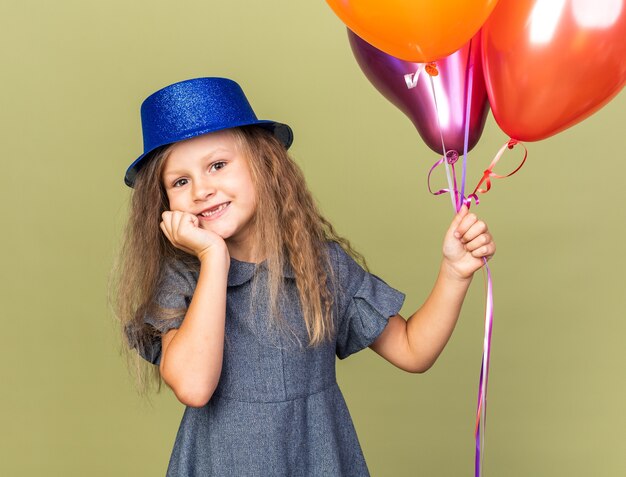 smiling little blonde girl with blue party hat holding helium balloons and putting hand on chin isolated on olive green wall with copy space