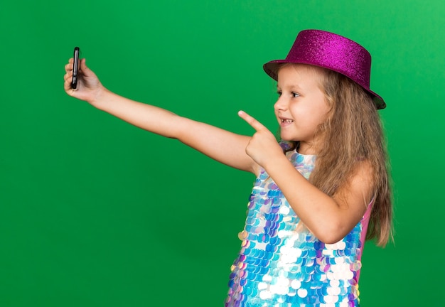 улыбающаяся маленькая блондинка с фиолетовой шляпой, держащая и указывающая на телефон, делающая селфи, изолированную на зеленой стене с копией пространства